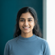 Headshot of a smiling young South Asian woman with a warm expression, against a soft deep teal and white background.